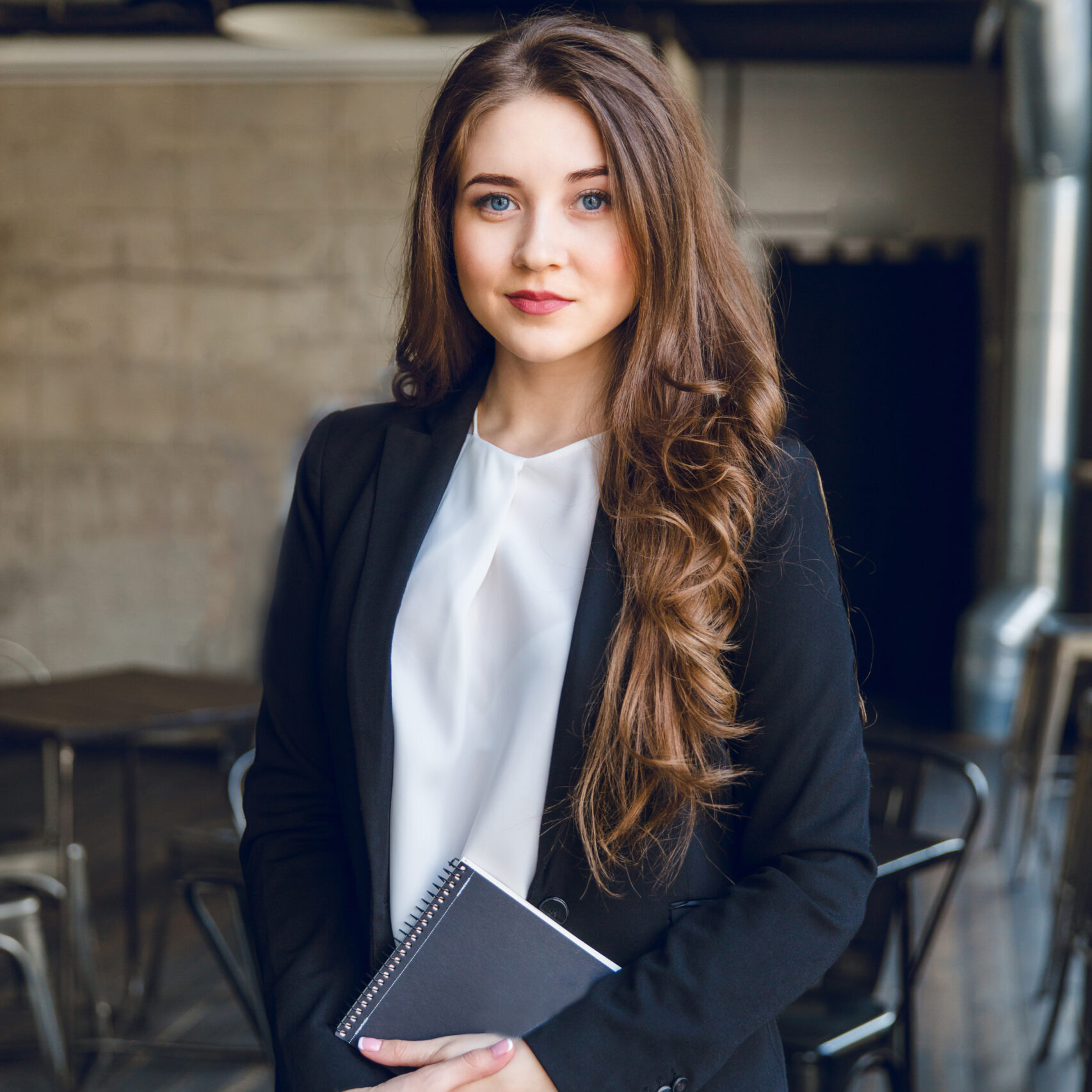 Brunette business woman with wavy long hair and blue eyes stands holding a notebook in hands. She wears a black suit and a white blouse. She is in a cafe with grey walls and dark furniture.